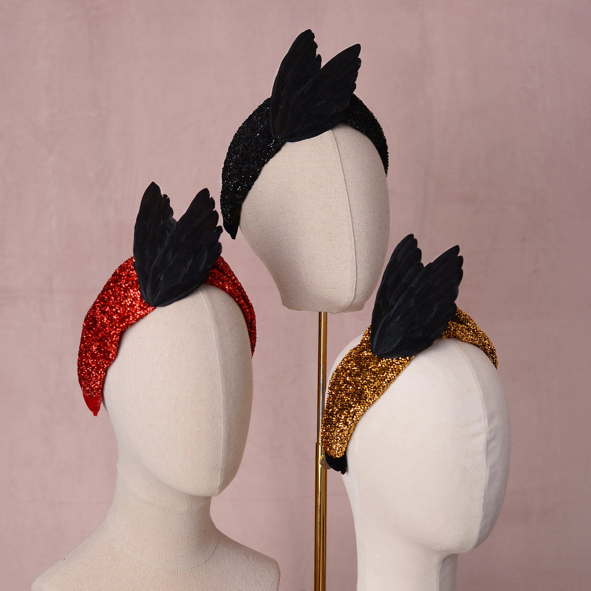 Three feathered headbands displayed on cream mannequin heads against a soft pink background. They are black, redor gold in a glittering fabric and adorned with a pair of black "bird wings "in an elegant v shape
