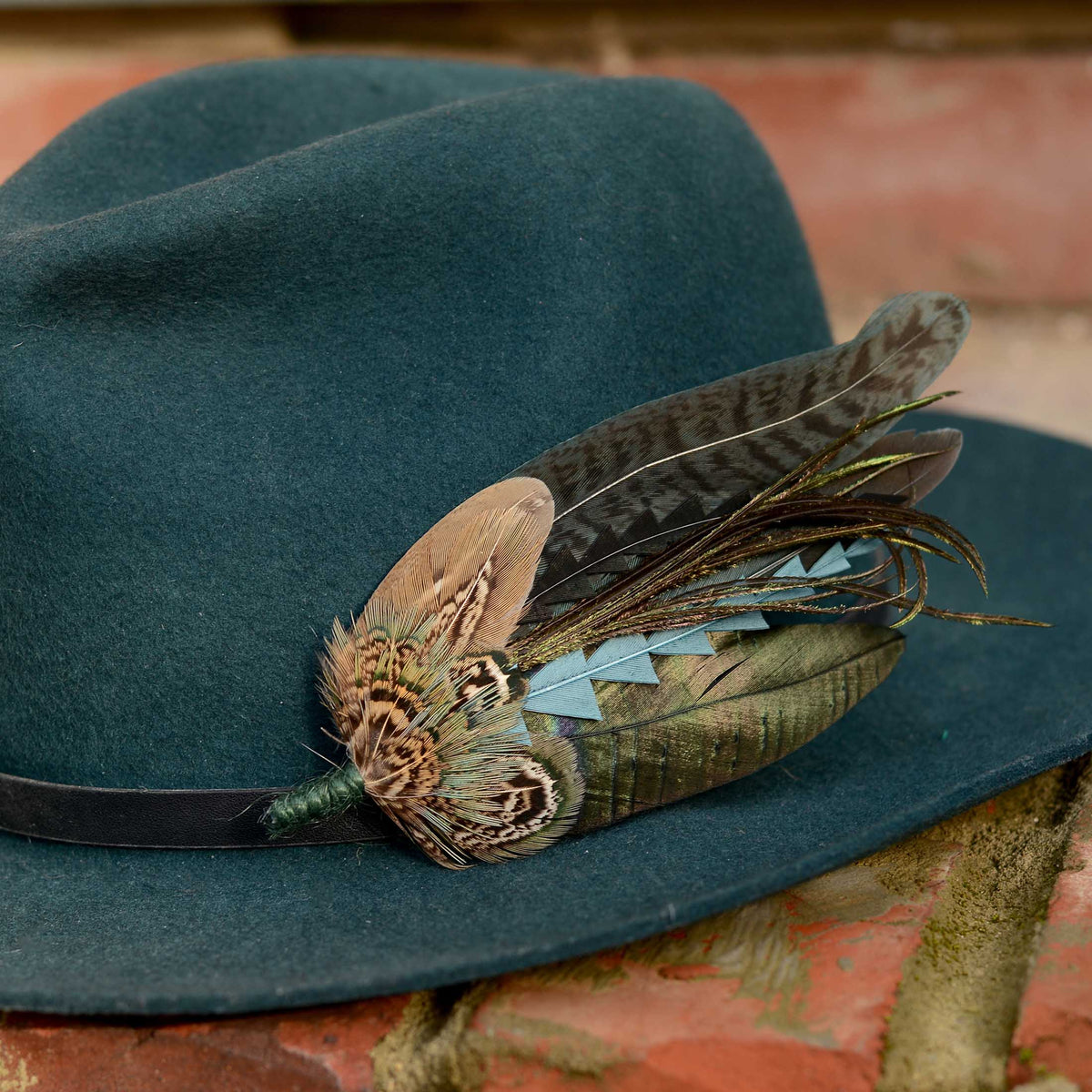 A dark teal felt hat with a black band, adorned with a large feather hat pin featuring black and teal-green feathers, resting on a rustic brick surface.