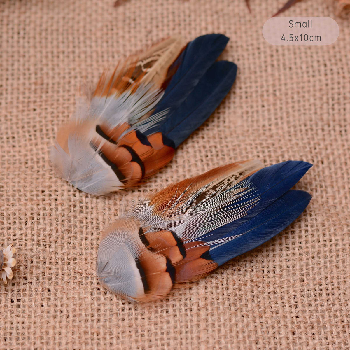 Two small decorative feather hair pieces with blue, brown, white, and black feathers, placed on a beige burlap background.