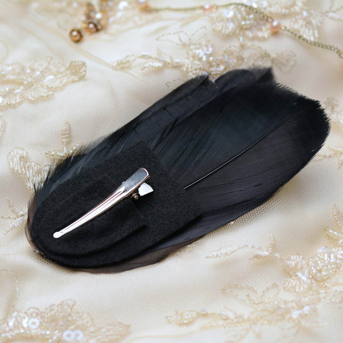 A black feather hair clip with a silver metal toned alligator style clip, displayed on a cream-colored embroidered fabric background.