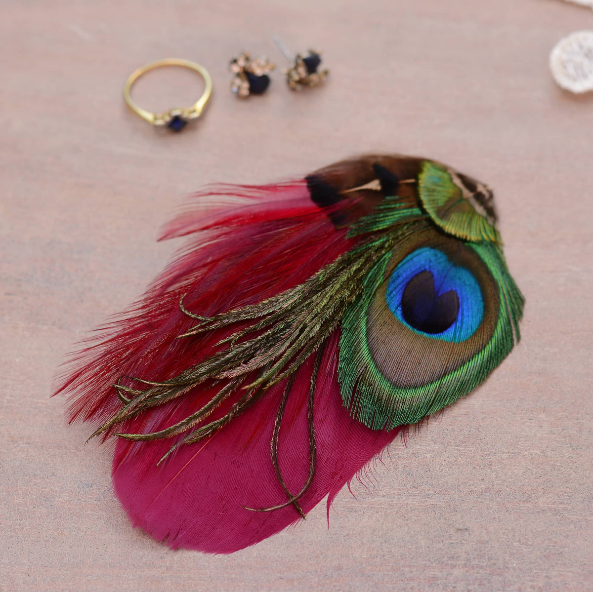 A decorative feather hair accessory featuring burgundy feathers with a peacock eye in green, blue, and brown, styled on a dusty-pink dressing table with small jewellery props in the background.