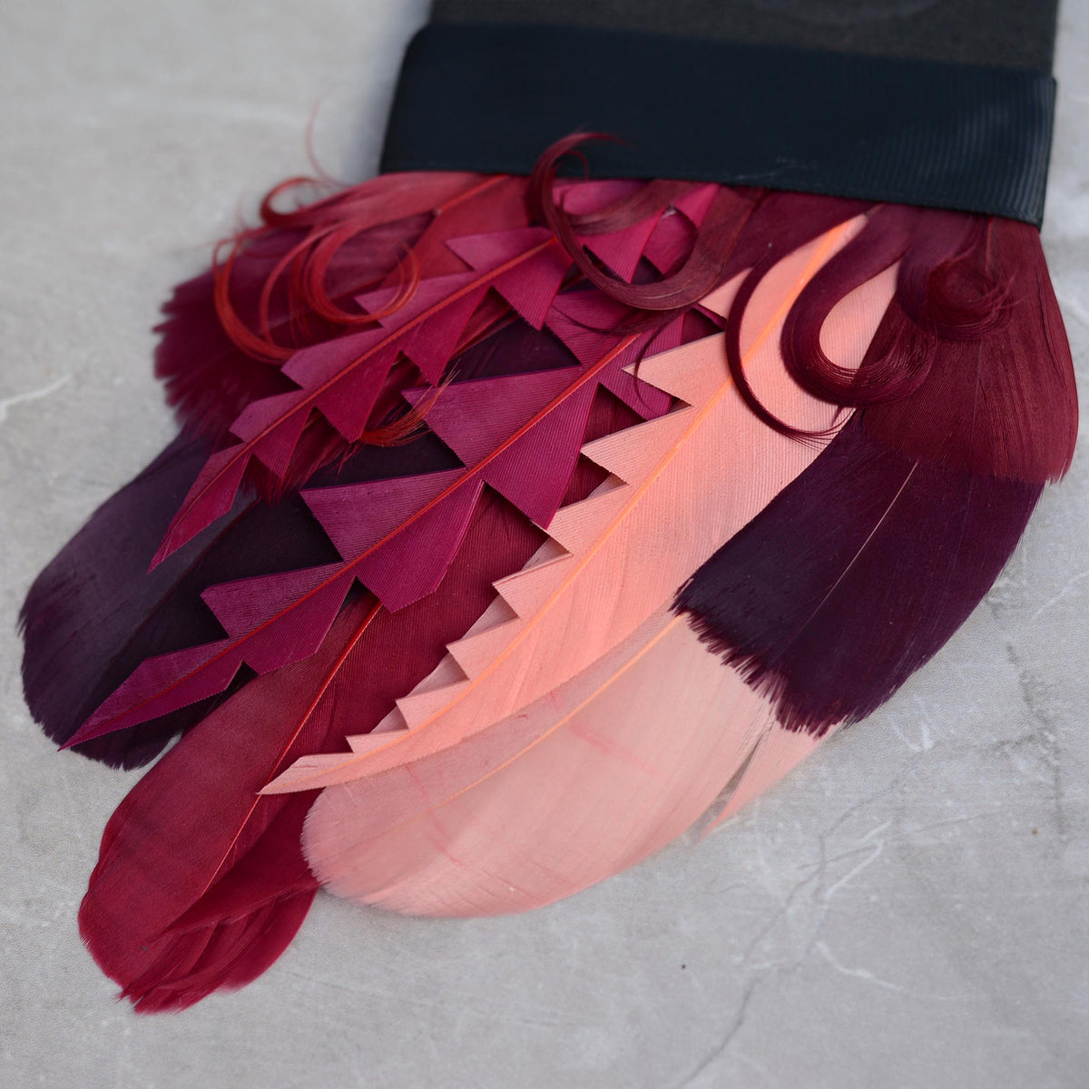 A close-up of a decorative feather suit accessory. A feather pocket square featuring layered goose feathers in deep burgundy, magenta, coral pink, and dark plum tones, cut into graphic shapes and curled for texture and depth.