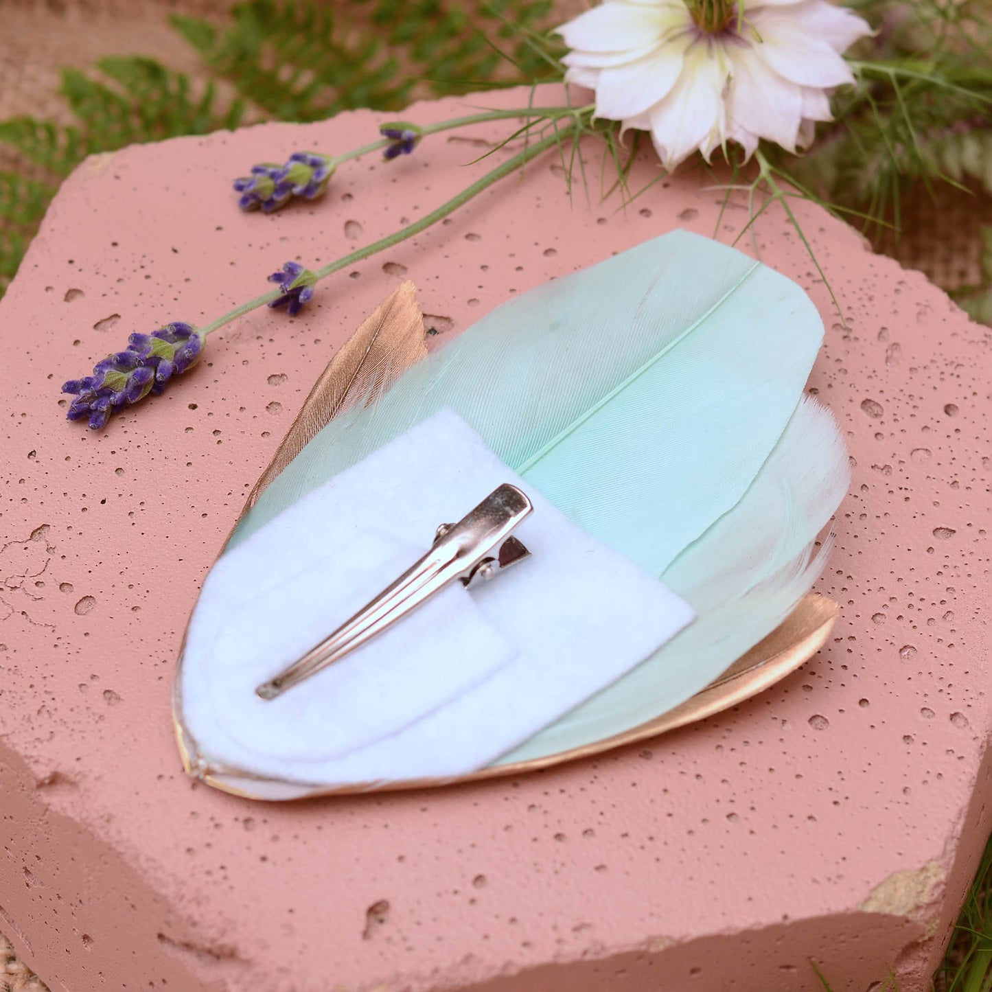 Large mint green and gold feather hair clip on a pink textured stone surrounded with burlap and flowers