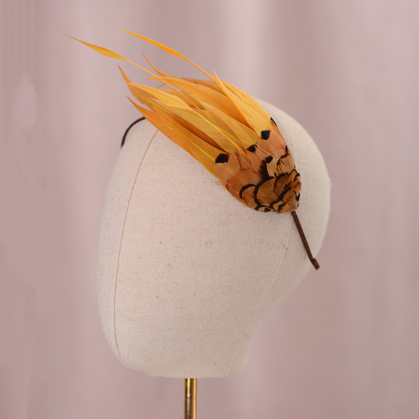 A decorative feather fascinator headpiece in yellow and orange-brown with black pattern details, displayed on a cream fabric mannequin head against a soft pink background.