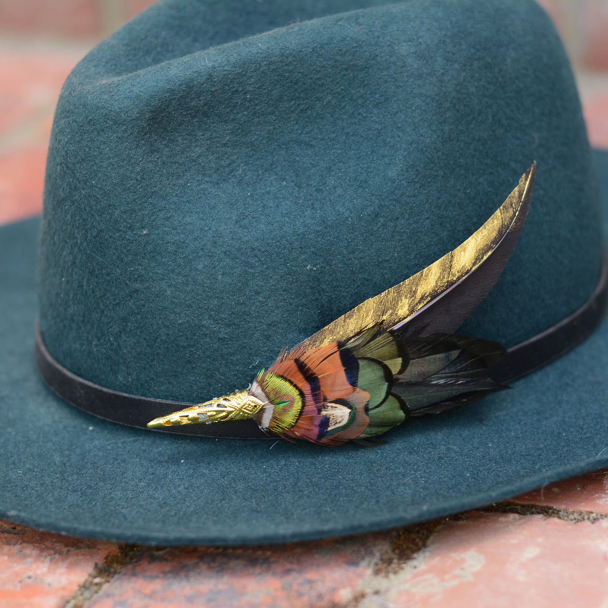 A dark teal felt hat with a black band and a decorative feather hat pin accent in gold, green, orange, and black, resting on a brick surface.