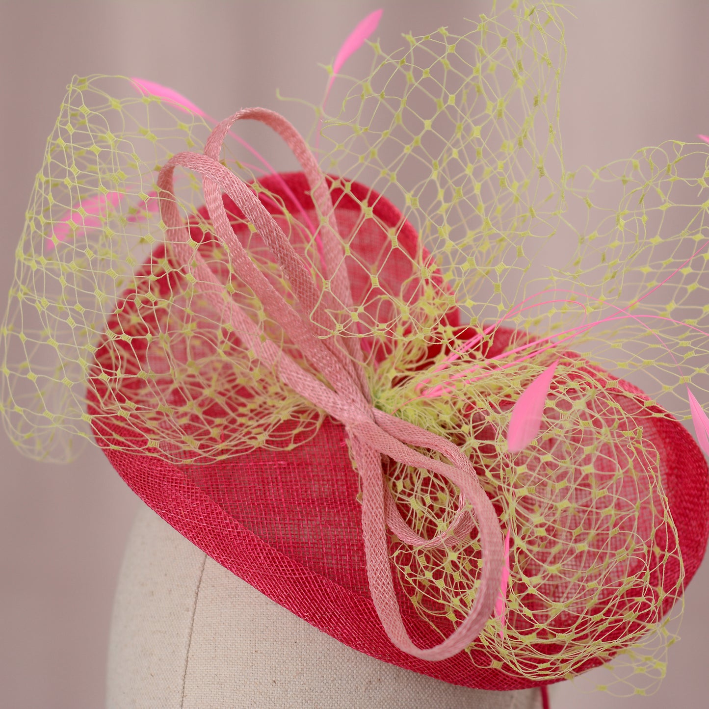 A bright fuchsia fascinator with yellow net veiling bow and pink feather details. The headpiece has a tonal pink sinamay bow. Ideal for weddings or formal events, this hat adds a pop of colour.