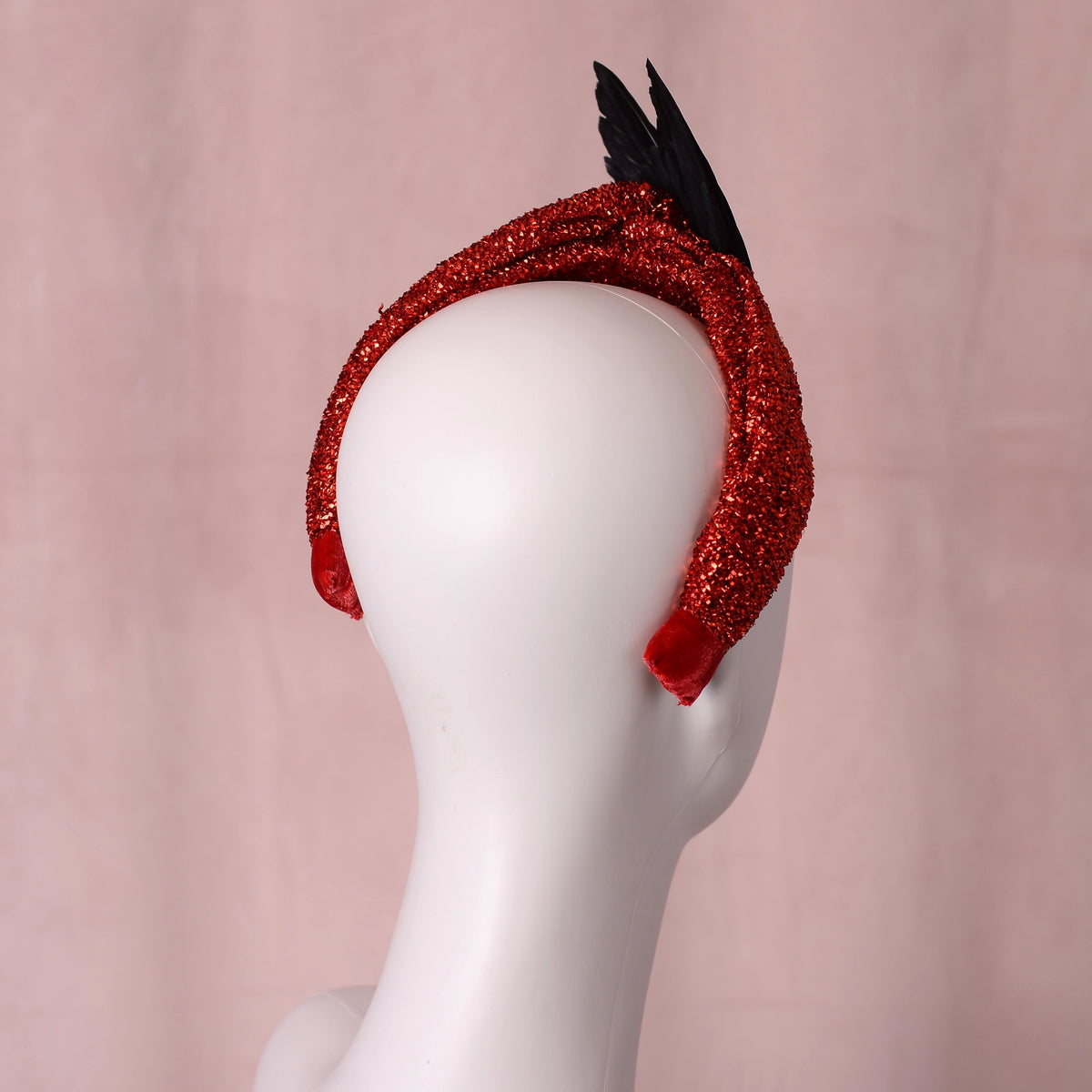 A red glitter fascinator headband with black feathers, displayed on a white mannequin head against a soft pink background.