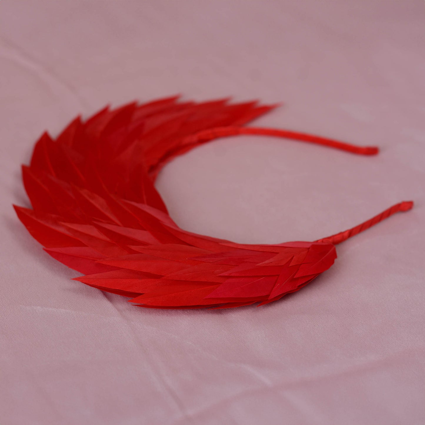 A bright red feather headband with layered, fan-like feathers on a thin red headband, displayed on a light pink fabric background.