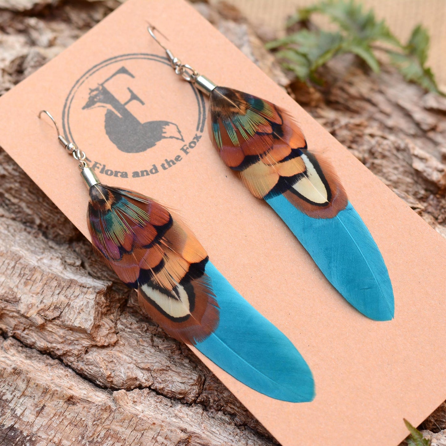 Long feather earrings in a vibrant teal turquoise layered with real male pheasant feathers in brown, copper and green On a logo-printed card on a bark background
