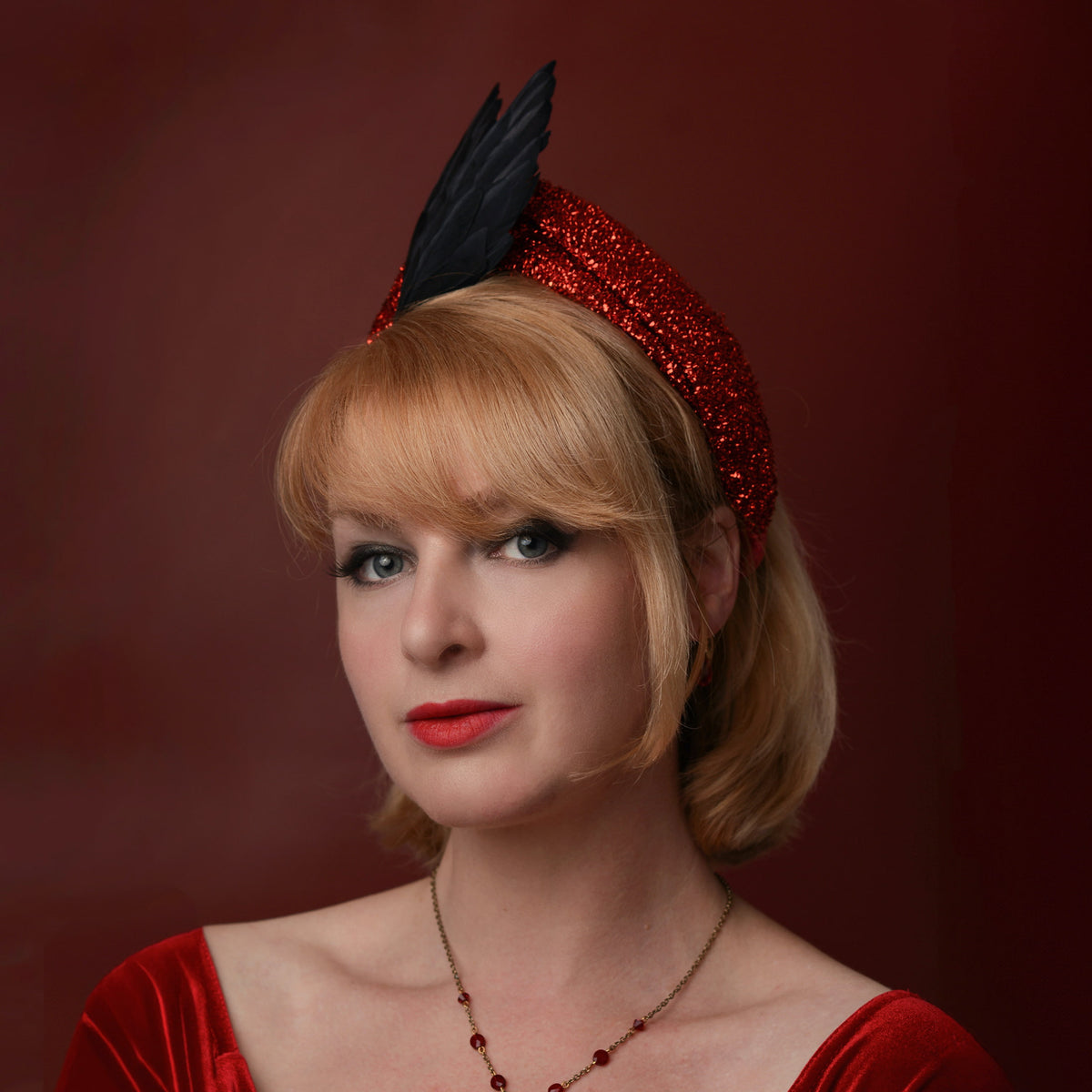 A woman with blonde hair wearing a red glitter headband with a black feather bird wing accent and a red velvet top, posed against a dark red background.