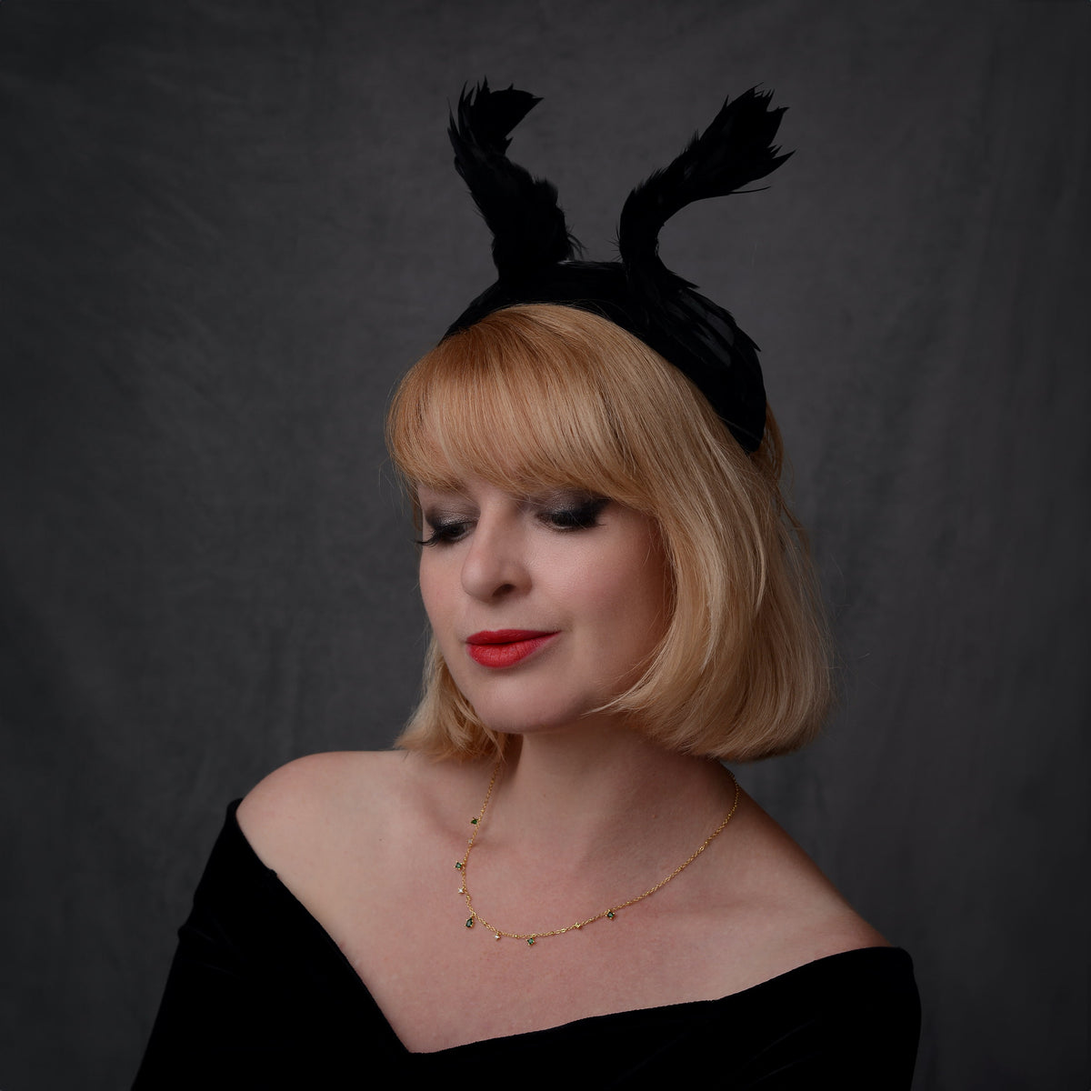 A woman with blonde hair wearing a winged black feather fascinator headband, a black off-the-shoulder top, and a delicate gold necklace, photographed against a dark gray studio backdrop. She has a witchy style.