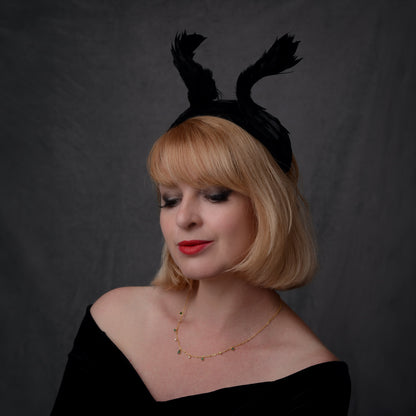 A woman with blonde hair wearing a winged black feather fascinator headband, a black off-the-shoulder top, and a delicate gold necklace, photographed against a dark gray studio backdrop. She has a witchy style.