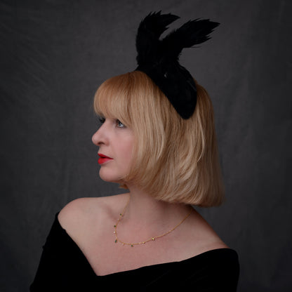 A woman with a short blonde bob and side-swept fringe is shown in profile  wears a black feather fascinator headpiece with wings sweeping backwards above the head, a black off-the-shoulder top, and a delicate gold necklace. She is against a dark gray studio background. 