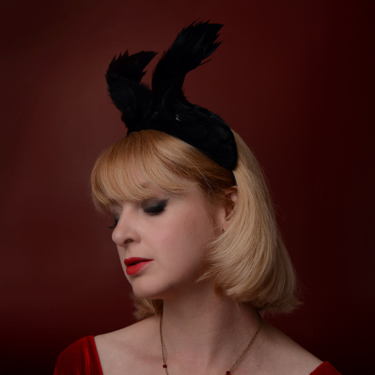 A blonde woman in profile wearing a gothic black feather winged fascinator headband, set against a dark red studio background.