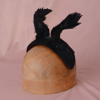 A black feathered headband fascinator displayed on a wooden hat block against a soft pink fabric backdrop.