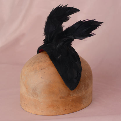 A black feathered fascinator hair accessory displayed on a wooden hat block against a soft pink fabric backdrop.