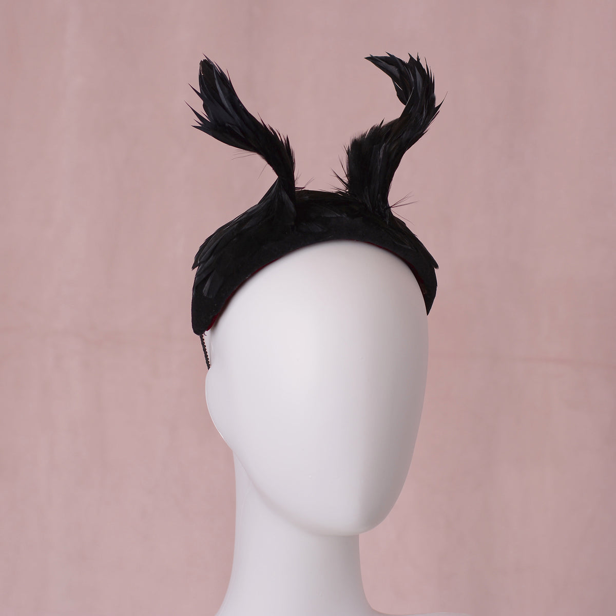 A black feather fascinator headpiece with twisted horns / wings that rise from the centre of the head displayed on a white mannequin head against a soft pink background.