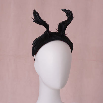 A black feather fascinator headpiece with twisted horns / wings that rise from the centre of the head displayed on a white mannequin head against a soft pink background.