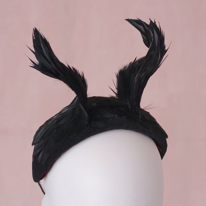 A  gothic black feather fascinator headpiece displayed on a white mannequin head against a soft pink background.