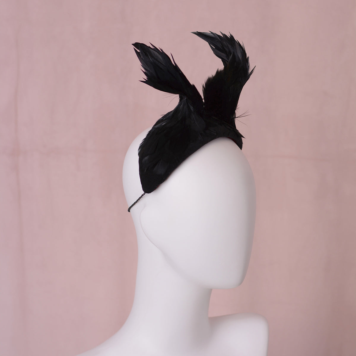 A black feather fascinator headpiece displayed on a white mannequin head against a soft pink background.