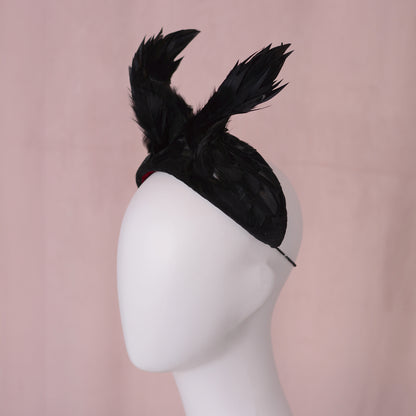 A black crow wing feather fascinator headpiece displayed on a white mannequin head against a soft pink background.