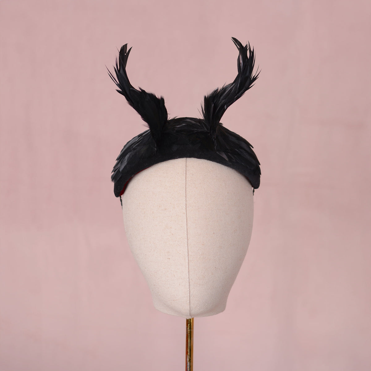 A black feather Halloween headpiece with two twisted feather horns, displayed on a cream fabric mannequin head against a soft pink background.