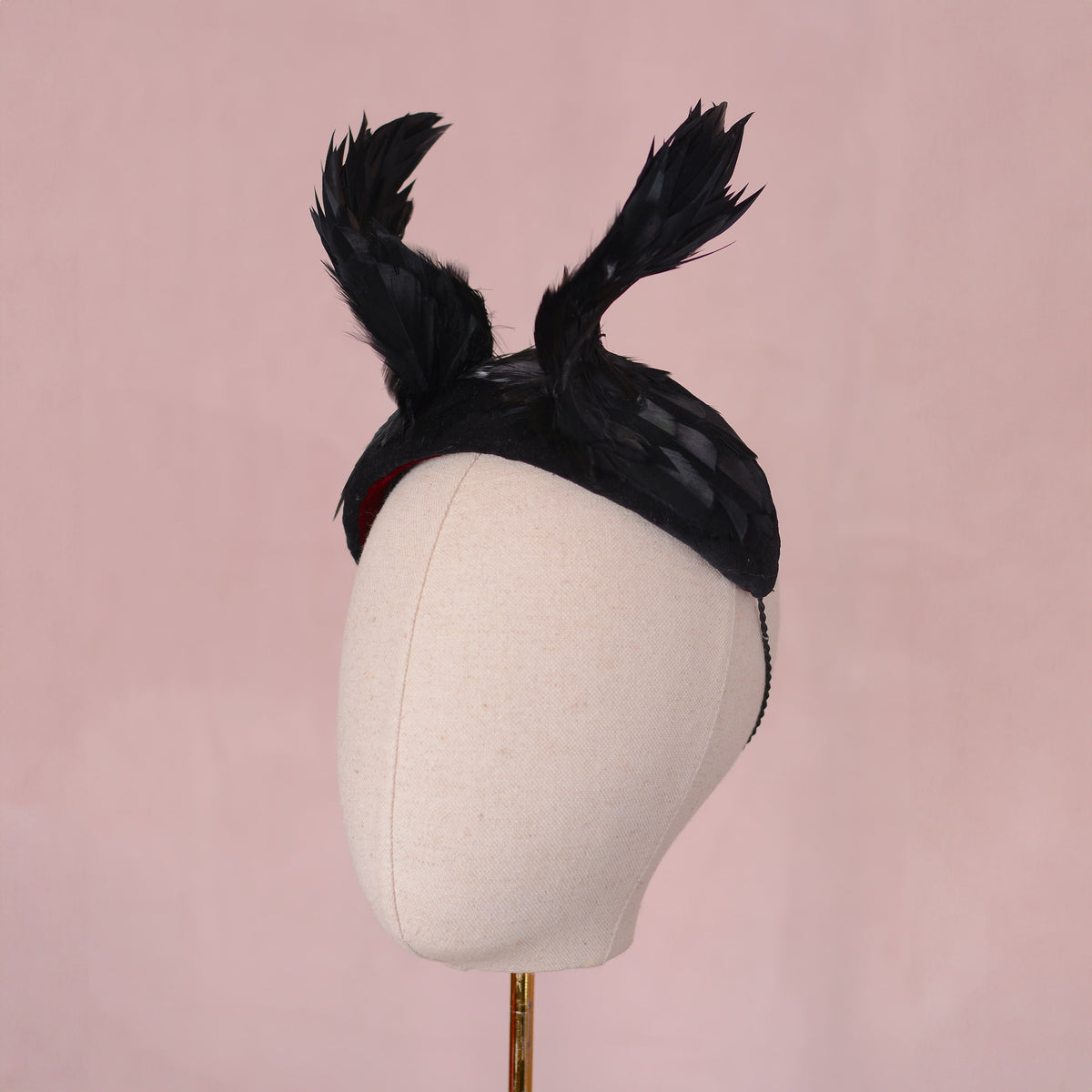 A black winged black feather headpiece displayed on a cream fabric mannequin head against a solid pink background. It has a witchy vibe ideal for glamorous halloween costumes.