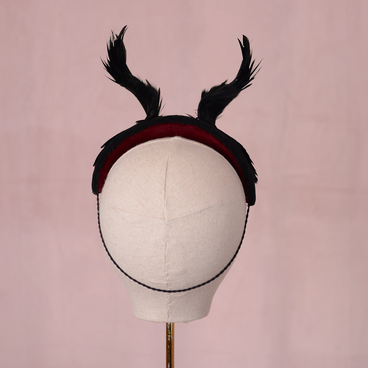 A black felt headband with two upright feathered horns and a dark red velvet lining, displayed on a cream fabric mannequin head against a soft pink background.