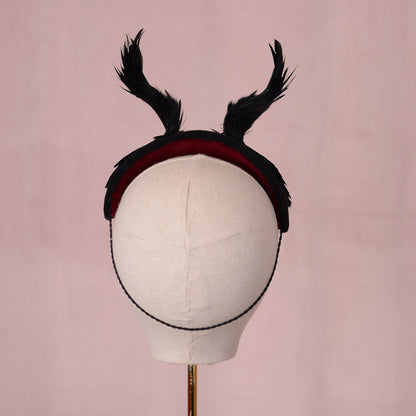 A black felt headband with two upright feathered horns and a dark red velvet lining, displayed on a cream fabric mannequin head against a soft pink background.