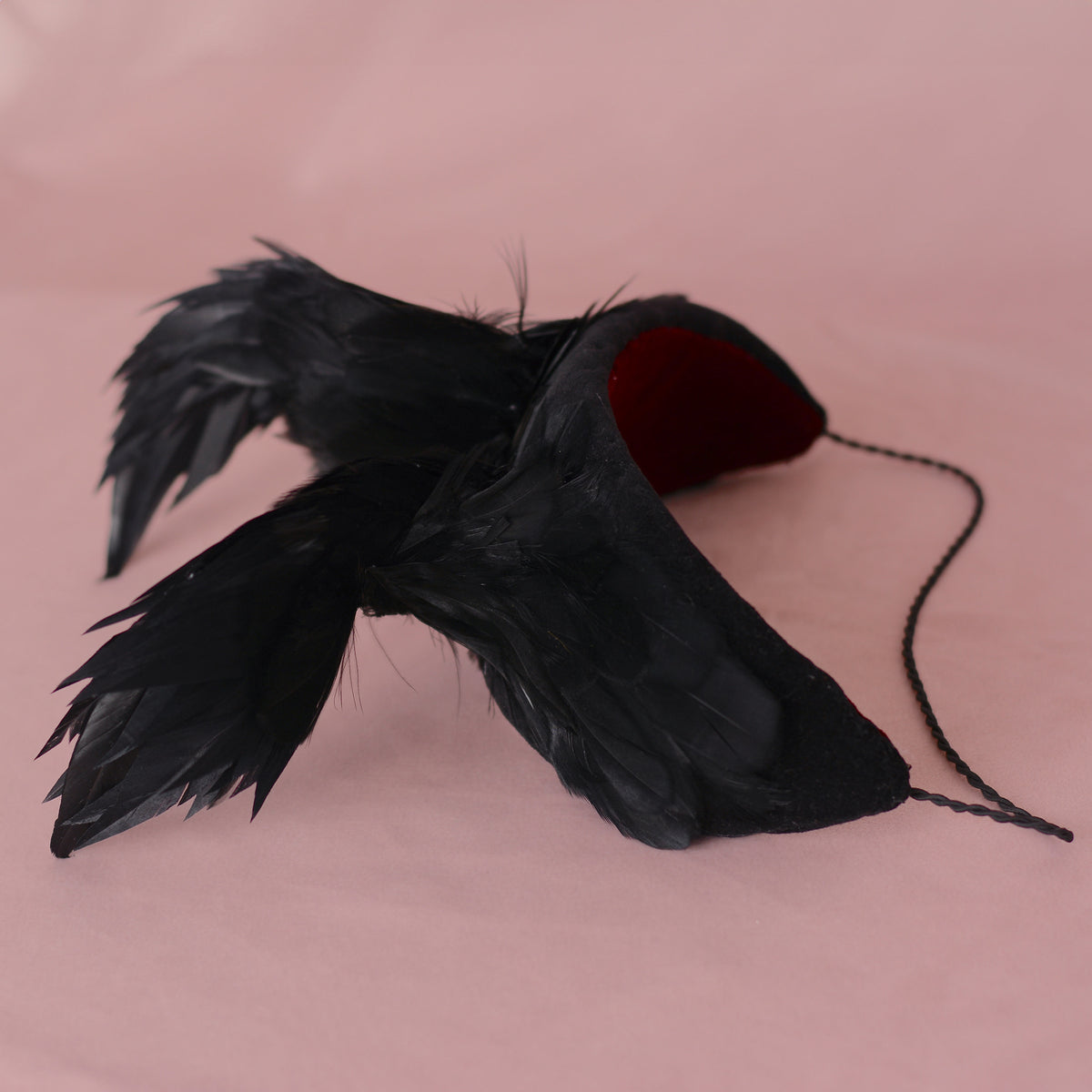 Black feathered headpiece with a red velvet inner lining and a black hat elastic, photographed on a solid pink background.