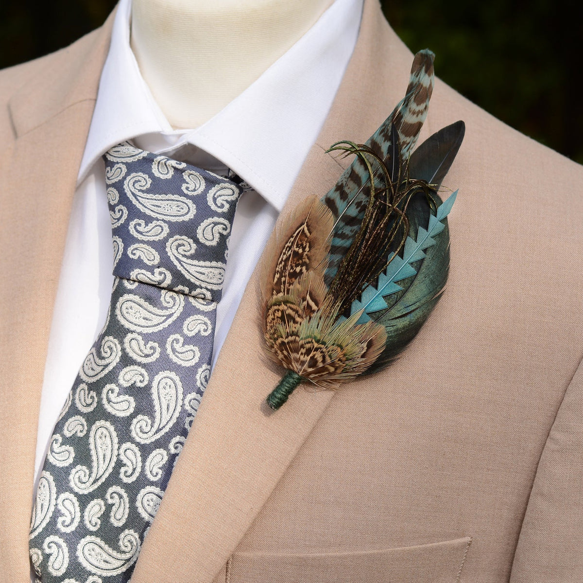 A close-up of a men's formal outfit featuring a beige suit jacket, white dress shirt, and a patterned blue-grey paisley tie, accessorised with a statement feather lapel pin. Ideal for a Groom's boutonniere