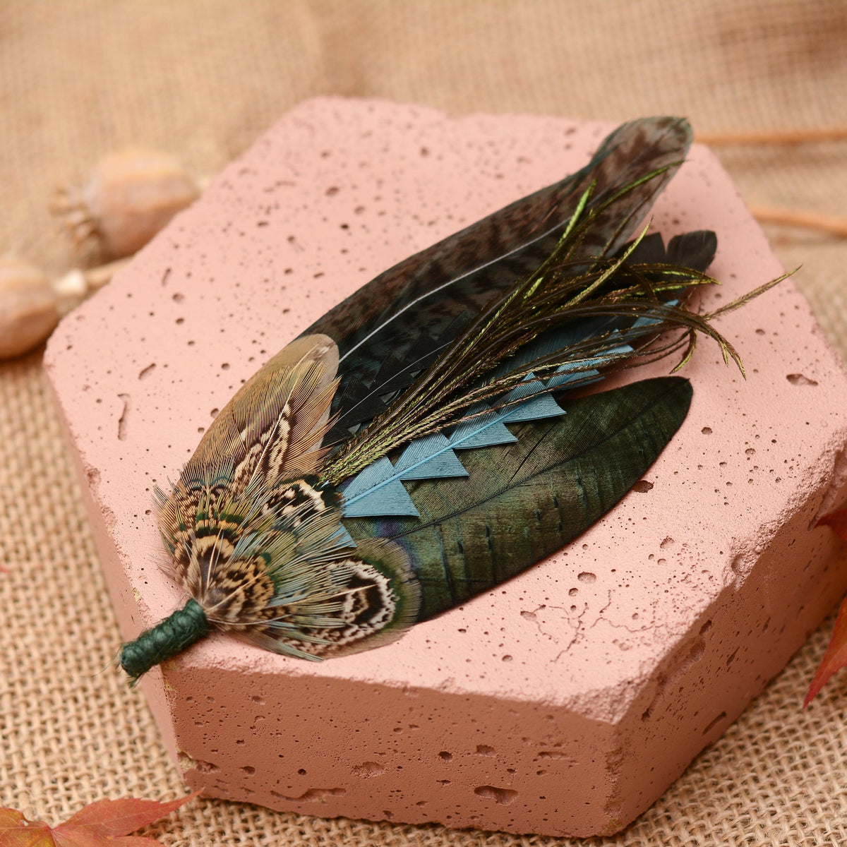 A decorative feather brooch made from a selection of natural feathers in green, blue, brown, and grey tones, wrapped with a green cord at one end, displayed on a pink stone block over a burlap surface.