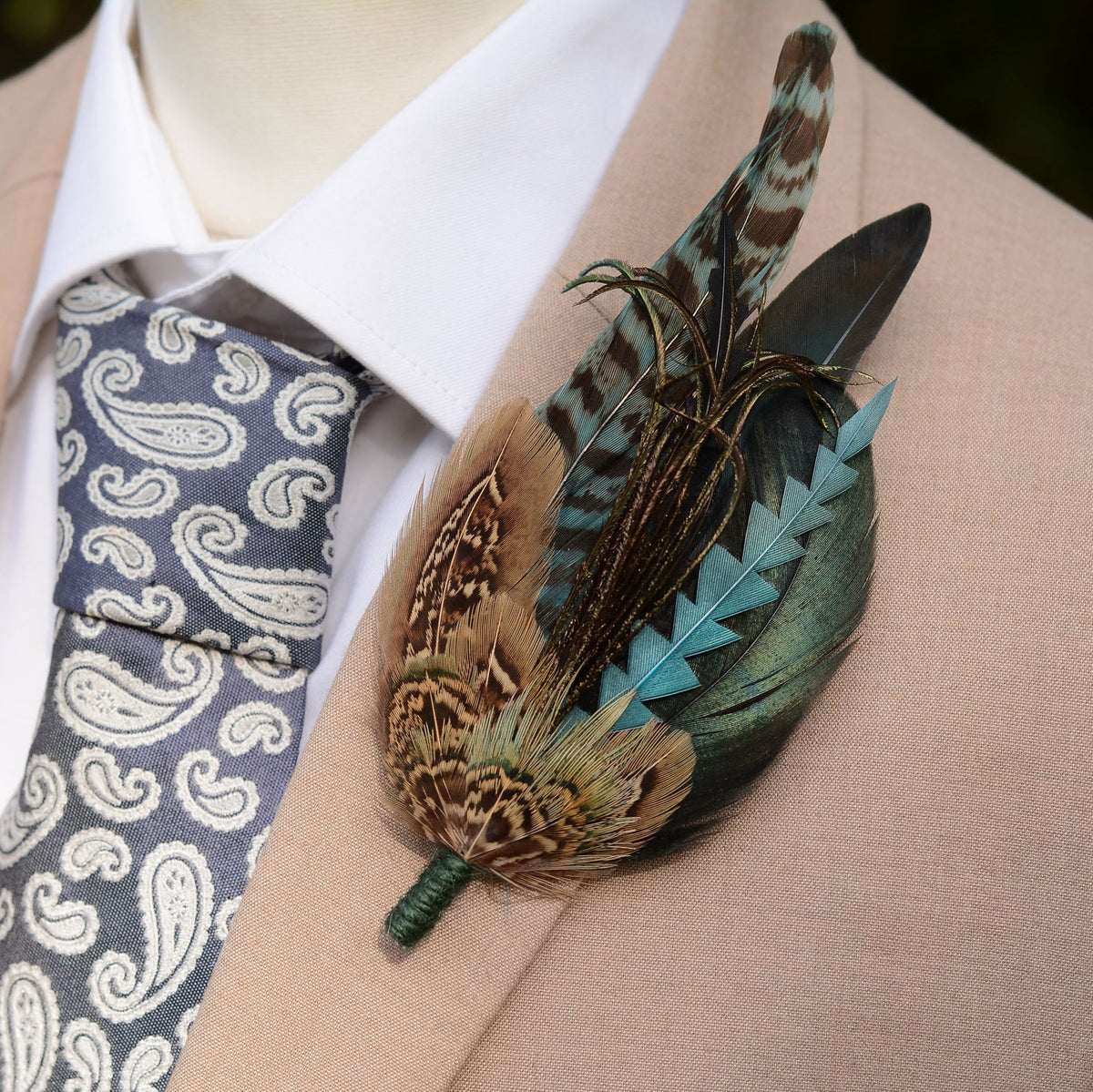 A large statment decorative feather boutonniere pin with brown, teal, and black feathers, pinned to a beige suit jacket over a white collared shirt and a grey paisley tie.