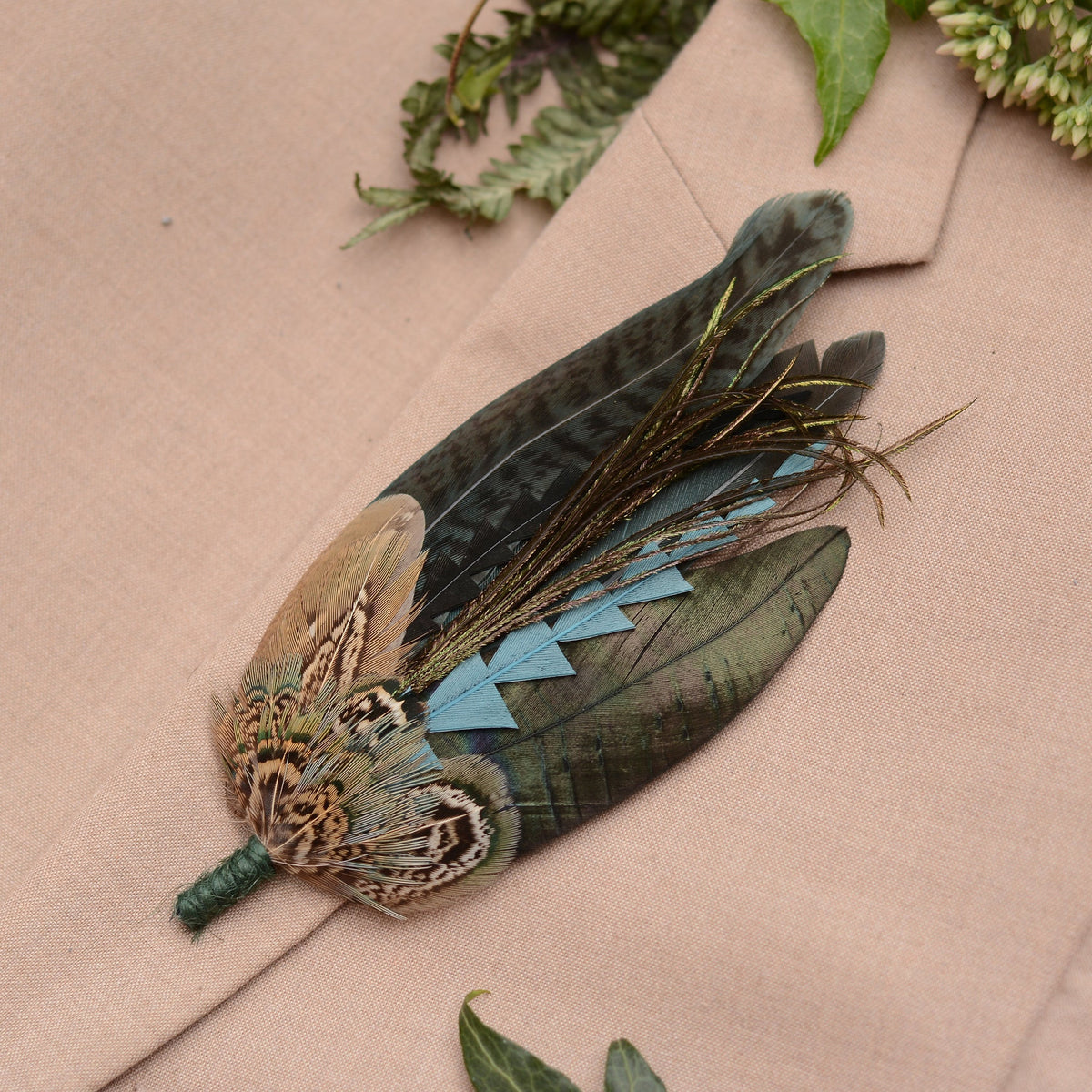 A decorative feather brooch pin accessory featuring a pheasant feathers and long iridescent feathers in green, blue, and brown tones, displayed on a beige fabric background with green leaves.