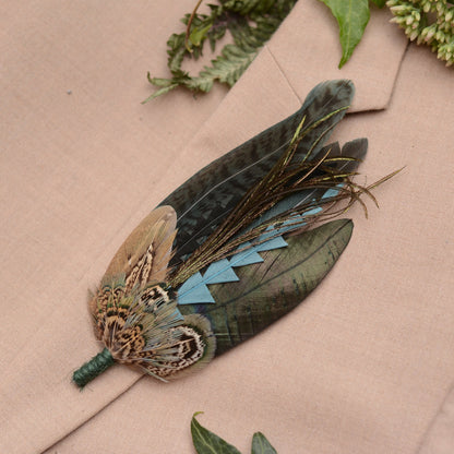 A decorative feather brooch pin accessory featuring a pheasant feathers and long iridescent feathers in green, blue, and brown tones, displayed on a beige fabric background with green leaves.