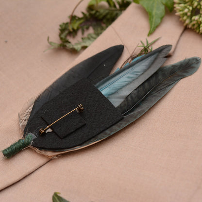 Feathered boutonniere  placed upside down on a beige suit jacket to show the felt backing and brooch pin with greenery in the background.