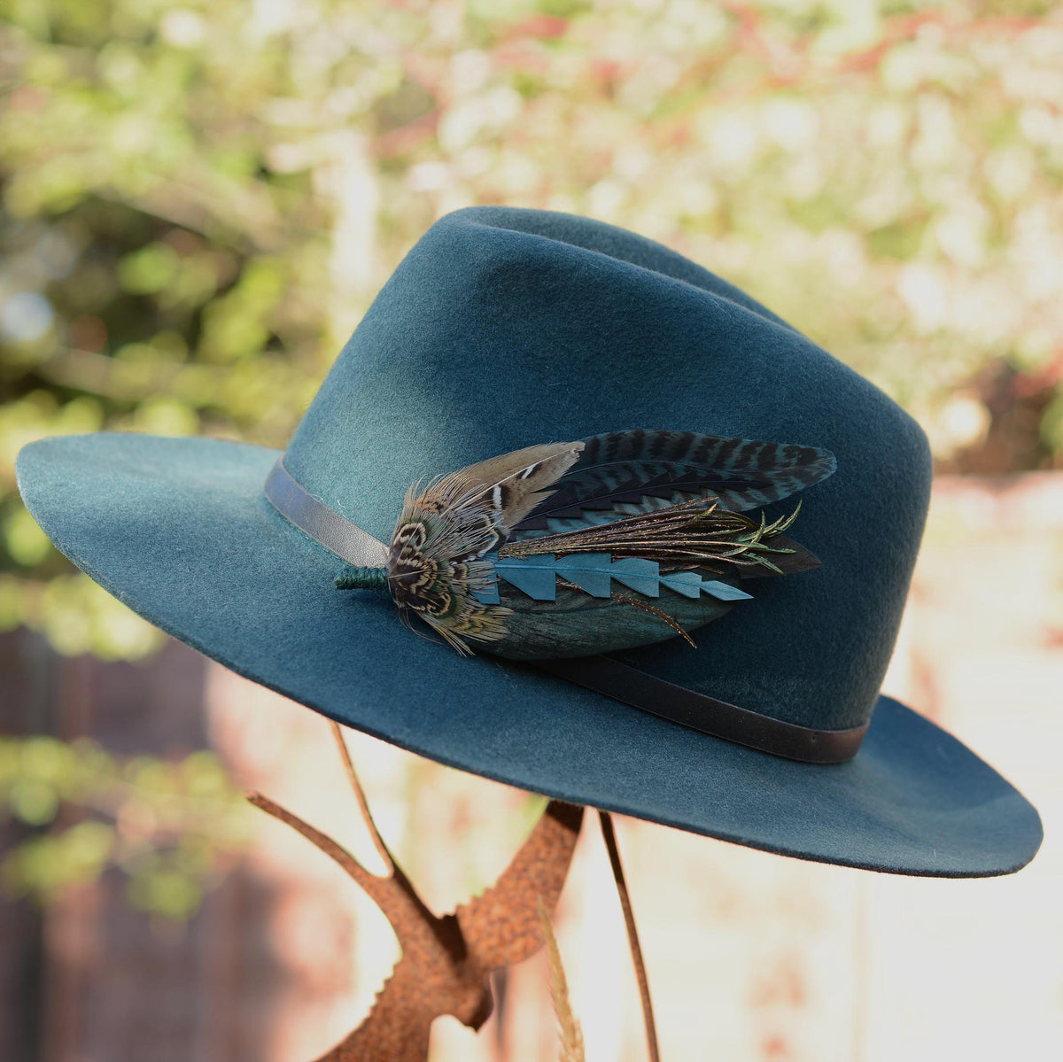 A teal blue felt cowboy hat with a dark brown band and a decorative feather pin, displayed on a rustic metal stand with a blurred outdoor garden background.