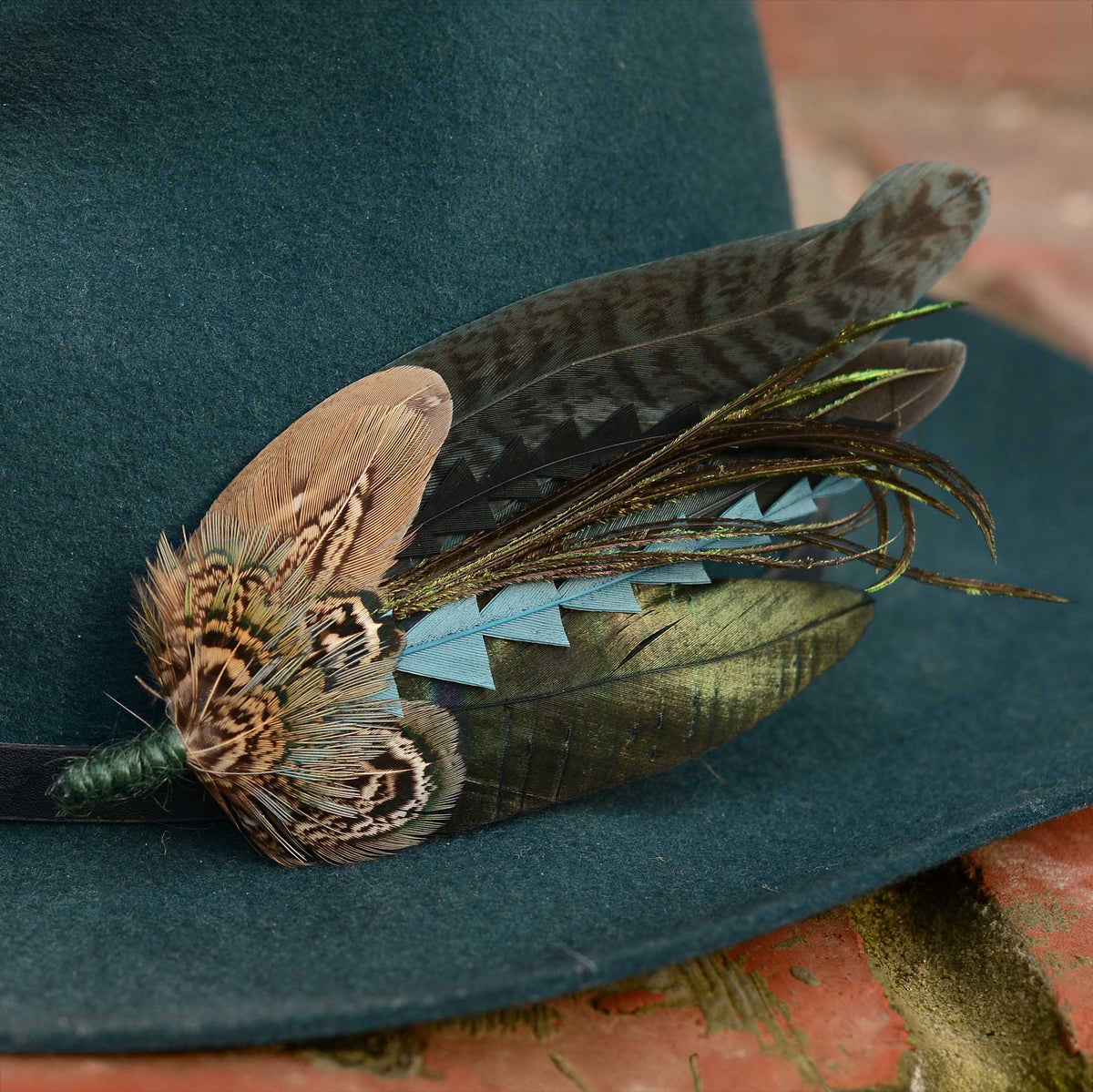 A dark teal felt hat featuring a decorative feather hat pin in teal, green and black resting on a brick surface.