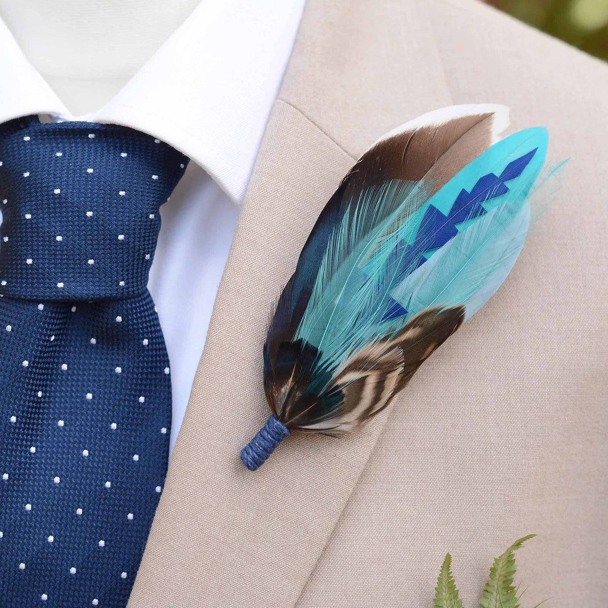 A decorative feather boutonniere pin with turquoise, blue, brown, and black feathers, pinned to a beige suit jacket worn with a blue polka-dot tie and white dress shirt.
