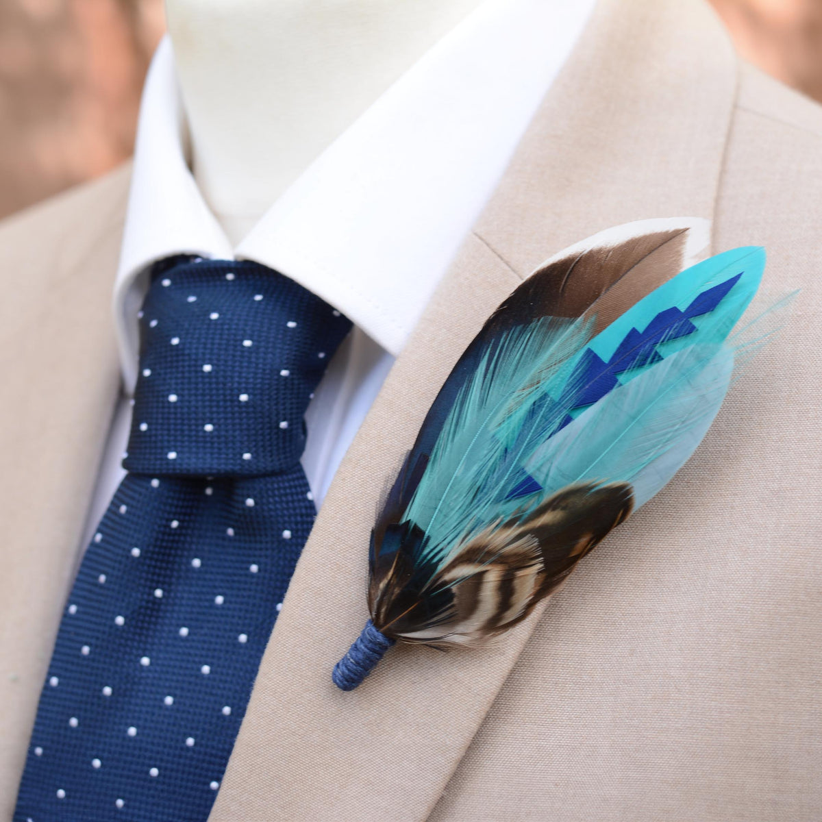 A close-up of a beige suit jacket styled with a white dress shirt, navy polka-dot tie, and a decorative feather boutonniere in turquoise, brown, and blue.