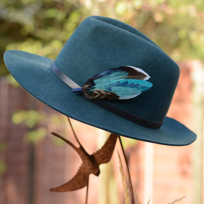 A teal felt wide-brim hat with a black band and a decorative blue feather hat pin, displayed outdoors on a metal stand with a blurred fence and foliage in the background.