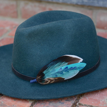 A dark teal felt fedora hat with a black band and a decorative feather hat pin in turquoise, brown and blue, resting on a red brick surface.