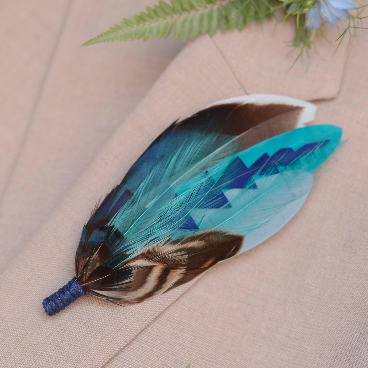 A decorative feather brooch pin with a blue twine wrapped base, featuring turquoise, blue, brown, black, and white patterned feathers, laid on a beige fabric surface.