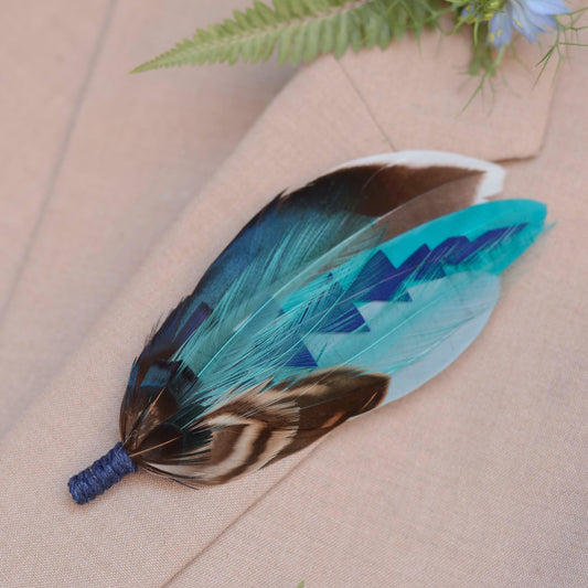 A decorative feather brooch pin with a blue twine wrapped base, featuring turquoise, blue, brown, black, and white patterned feathers, laid on a beige fabric surface.