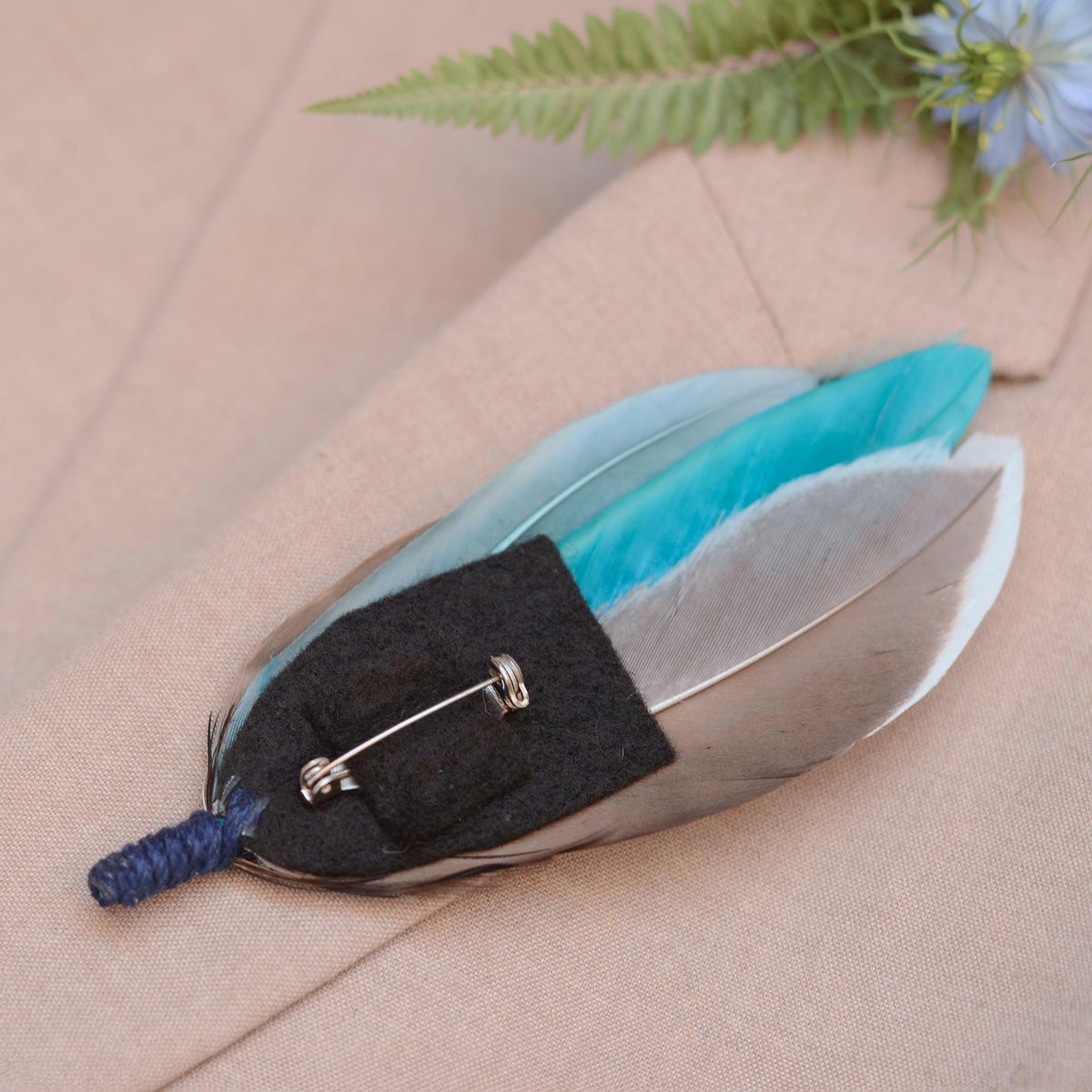 A decorative feather pin with a turquoise blue feathers on a black felt base with a silver safety pin, resting on a beige fabric background.