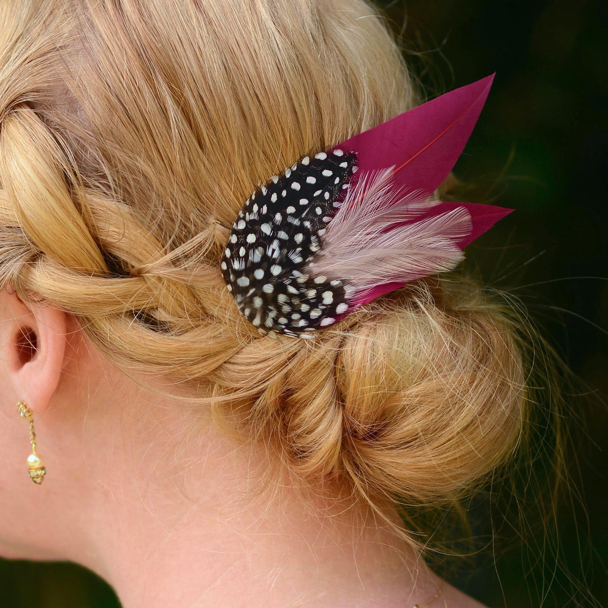 A close-up of a blonde woman's hair styled in a low braid with a decorative hair accessory featuring burgundy and blush pink feathers, and black-and-white polka dot feathers.