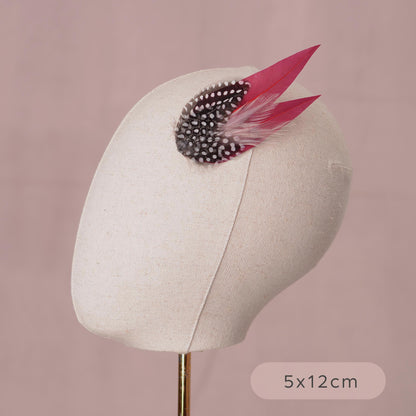 A decorative  feather hair accessory displayed on a fabric mannequin head against a soft pink background. The accessory features two burgundy feathers layered with wispy blush pink feathers and polka dot guinea fowl feathers.