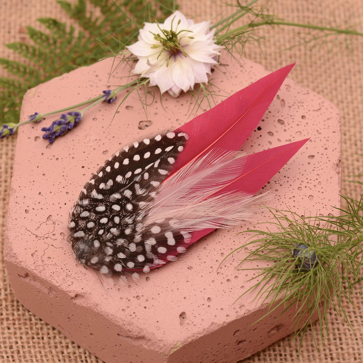A decorative feather hair accessory displayed on a pink hexagonal block, featuring burgundy feathers layered with fine and delicate blush pink feathers and black feathers with white polka dot spots, styled with flowers and greenery on a burlap background.