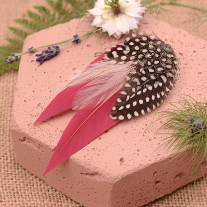 A decorative feather hair accessory displayed on a pink hexagonal block, featuring burgundy feathers layered with fine and delicate blush pink feathers and black feathers with white polka dot spots, styled with flowers and greenery on a burlap background.