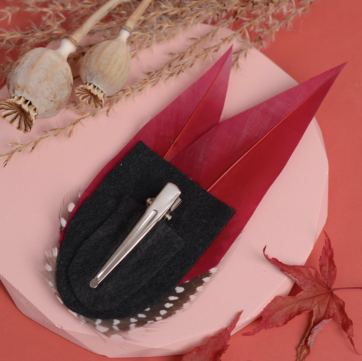 A decorative feather hair clip accessory with a black and white spotted feathers and burgundy and blush pink feathers, shown upside down displaying a black felt base and silver toned alligator style clip styled on a pink surface with dried botanical props and a red leaf.